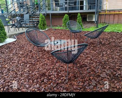 Sessel aus Rattan in einem Stadtpark. Schwarzer Drahtsessel auf einem dekorativen braunen Chip. Gartenmöbel. Ein Ort zum Entspannen in der Nähe eines Cafés in einem Stockfoto