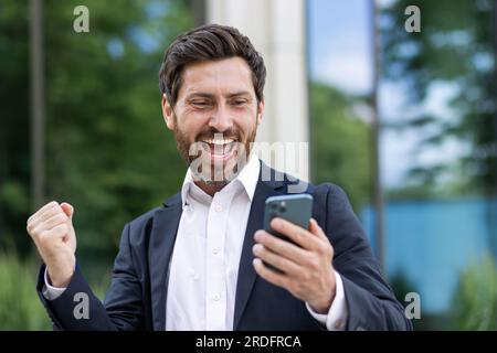 Ein erfahrener Geschäftsmann mit Telefon feiert den Sieg, der Chef im Geschäftsanzug erhielt eine gute Benachrichtigung online, ein Mann außerhalb des Bürogebäudes hält die Gewinnergeste Hand hoch. Stockfoto