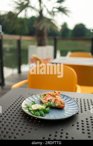 Croissant-Sandwich mit Lachs, pochiertem Ei und Gurke auf dem Teller. Frühstück im Café oder Café. Lifestyles. Stockfoto