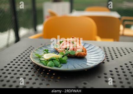 Croissant-Sandwich mit Lachs, pochiertem Ei und Gurke auf dem Teller. Frühstück im Café oder Café. Lifestyles. Stockfoto