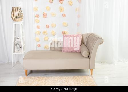 Beigefarbenes klassisches, aber modernes Sofa im Himmelsstil mit pinkfarbenen Kissen und 3D Pfingstrosen zur Wanddekoration in weißem Innenraum. Stockfoto