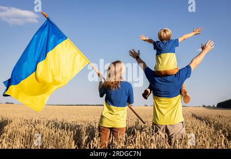 Glückliche ukrainische Familie in gelb-blauen T-Shirts steht zwischen Stacheln im Weizenfeld, Kind sitzt auf Dads Schultern, Frau hält die Flagge der Ukraine. nati Stockfoto