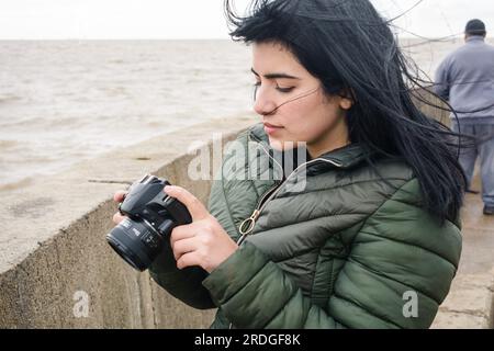 Junge lateinische Touristin venezolanischer ethnischer Herkunft eine junge Frau im Freien, die auf dem Pier steht und auf den Bildschirm der Digitalkamera schaut und die Fotos betrachtet Stockfoto