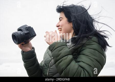 Junge lateinin venezolanischer Herkunft, die mit ihrer Digitalkamera glücklich lächelt und Inhalte für ihre sozialen Netzwerke im Freien in einer Kälte und Wolke erstellt Stockfoto