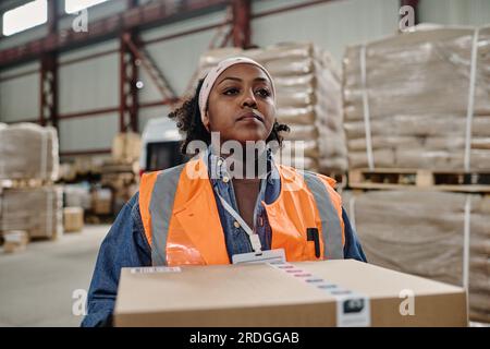 Eine afroamerikanische Mitarbeiterin, die während ihrer Arbeit im Lagerhaus eine Kiste zur Auslieferung trägt Stockfoto