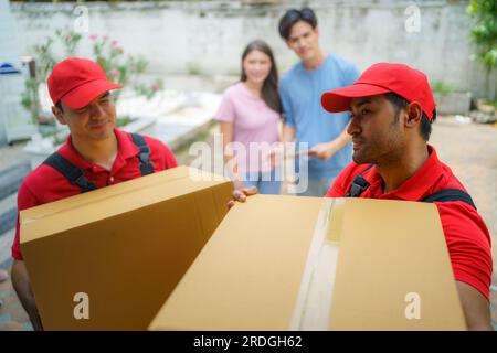 Ein asiatisches Paar steht zusammen und beobachtet mit Vorfreude, wie Arbeiter in ihr neues Zuhause ziehen. Spannung füllt die Luft, wie sie eifrig erwarten Stockfoto