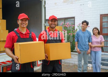 Ein asiatisches Paar steht zusammen und beobachtet mit Vorfreude, wie Arbeiter in ihr neues Zuhause ziehen. Spannung füllt die Luft, wie sie eifrig erwarten Stockfoto