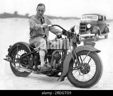 Daytona Beach, Florida: 13. Februar 1935 der britische Rennfahrer Captain Malcolm Campbell probiert die Harley Davidson eines Polizisten aus, aber er wird nicht annähernd die 300 km/h erreichen, die er in seinem Bluebird Rennwagen am Strand erreichen möchte. Stockfoto