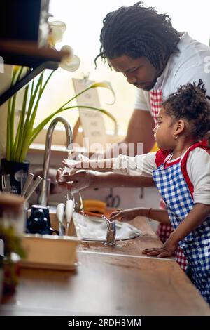 Junger Vater wäscht mit ihrer kleinen Tochter in einer Hausküche Geschirr ab, arbeitet als Team. Zuhause, Familie, Lifestyle-Konzept. Stockfoto