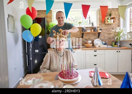 Ältere Frau feiert ihren Geburtstag, sitzt am Tisch vor einem Geburtstagskuchen mit Kerzen. Ihr Sohn steht hinter ihr und arrangiert ihre cr Stockfoto