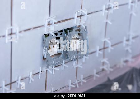 Ein Porträt von zwei angeschlossenen elektrischen Wandsteckdosen ohne Schutzkappen aus Kunststoff in einer neu gefliesten, unfertigen Wand, immer noch mit weißem Kunststoff Stockfoto
