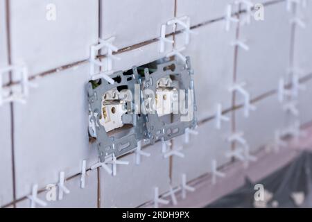 Nahaufnahme von zwei angeschlossenen Steckdosen ohne Schutzkappen in einer neu gefliesten, unfertigen Wand, immer noch mit weißem Plastik Stockfoto