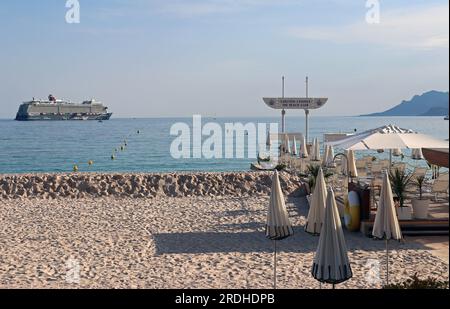 Am frühen Abend fällt die Sonne auf einen Teil des Carlton Beach Club in Cannes, bevor die Saison beginnt. Draußen auf dem Meer liegt ein riesiges TUI-Kreuzfahrtschiff vor Anker. Stockfoto