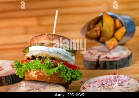 Saftiger Burger mit gesalzenem Fleisch und Ei auf einer Holzplatte mit Salzstücken an der Seite und gebackenen Kartoffeln im Hintergrund Stockfoto