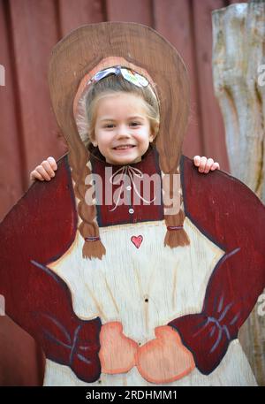 Das kleine Mädchen posiert hinter einer Holzfigur der Frau eines Bauern. Sie stockt ihren Kopf durch die Öffnung und lächelt. Sie besucht ein Erntefest Stockfoto