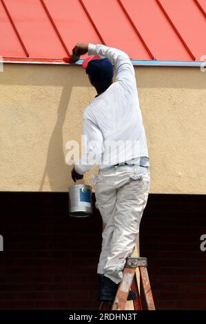 Der Mann steht auf der obersten Stufe der Leiter, während er die Dachlinie am Bürogebäude in der Innenstadt von Albuquerque, New Mexico, lackiert. Er hält eine Farbdose und Sprea in der Hand Stockfoto