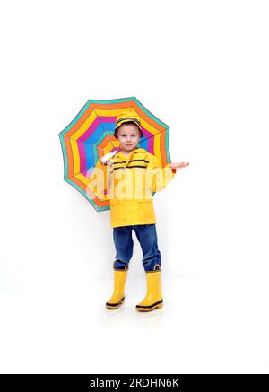 Little boy Kontrollen für Regen, während in einem gelben Slicker und Gummistiefel und Hut bekleidet. Er ist ein multi-gestreifte Schirm in einem weißen Raum. Stockfoto