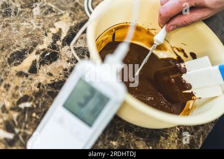 Die Hände des männlichen Küchenchefs messen die Temperatur von geschmolzener dunkler Schokolade mit einem professionellen Süßwarenthermometer. Nahaufnahme eines professionellen Konditors bei der Arbeit. Anzeigenkonzept für Thermometer. Hochwertiges Foto Stockfoto