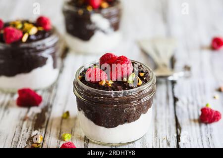 Dunkler Schokoladen-Chia-Pudding mit Himbeeren und Pistazien oben in einem Glasgefäß auf weißem Holzhintergrund. Serviert mit Kokosnuss-Schlagsahne. Stockfoto