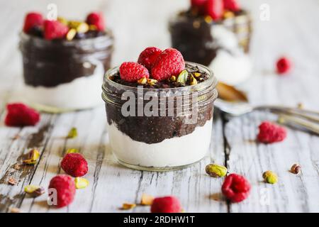 Dunkler Schokoladen-Chia-Pudding mit Himbeeren und Pistazien oben in einem Glasgefäß auf weißem Holzhintergrund. Serviert mit Kokosnuss-Schlagsahne. Stockfoto