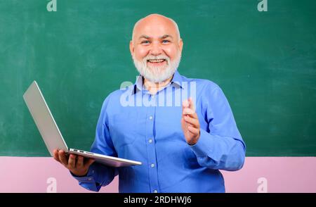 Lächelnder Lehrer mit Laptop vor der Tafel im Klassenzimmer. Lehrer, der Schülern Unterricht gibt. E-Learning an der Universität. Mit männlichem Lehrer Stockfoto