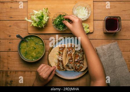 Die Hand einer Frau bereitet einen Taco aus mariniertem Fleisch zu. Teller mit Tacos, Sauce und Gemüse auf einem Holztisch. Draufsicht. Stockfoto