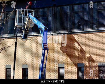 Gründliche Fensterreinigung. Hochdruckwasserstrahl. Hydraulischer Arbeitskorb und Plattform am Teleskopausleger. Backsteinaußenwand. Arbeiter und Wasserschlauch Stockfoto