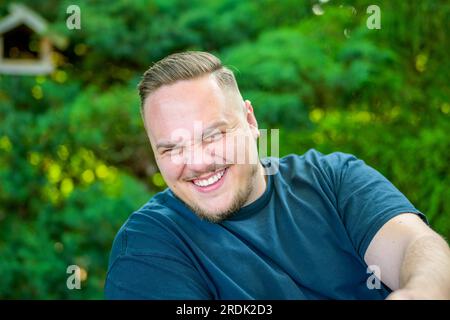 Porträt eines jungen Mannes mit Bart, der im Garten sitzt und mit einem frechen und begeisterten Lächeln in die Kamera schaut Stockfoto