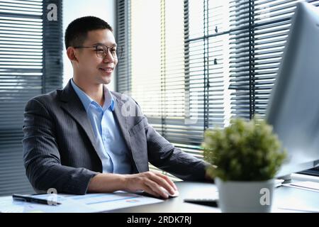 Eleganter asiatischer Geschäftsmann mit Brille, der auf einen Computermonitor schaut und Online-Datenmarktanalysen am modernen Arbeitsplatz durchführt Stockfoto