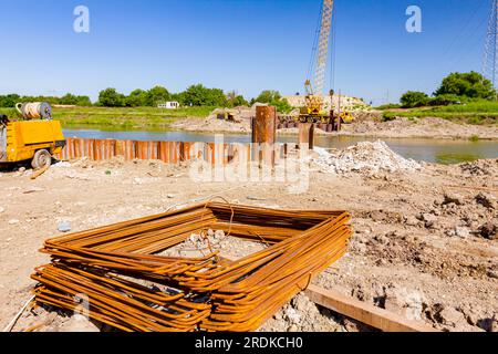 Rostige quadratische Bewehrungsstäbe auf der Baustelle. Am Flussufer sind Metallpfähle für das Brückenfundament installiert, im Hintergrund ist der Kranwitze Stockfoto