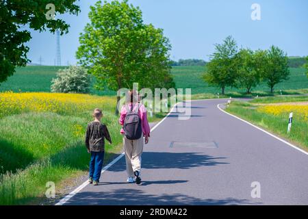 Mama und Sohn wandern an einem sonnigen Tag auf einer Landstraße zwischen Feldern mit blühenden Rapssamen. Eine Frau und ein Kind mit Rucksäcken laufen am Morgen die Straße entlang Stockfoto