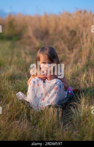 Ein wunderschönes kleines Mädchen sitzt im Gras auf der Wiese. Portrait eines hübschen, kleinen Mädchens auf dem Sommerfeld. Das durchdringende und ernste Aussehen des B Stockfoto