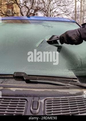 Autofenster im Winter reinigen. Der Mensch kratzt mit einem Plastikkratzer von der Windschutzscheibe eines blauen Autos. Hand in schwarzem Lederhandschuh im CO Stockfoto