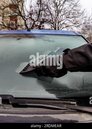 Autofenster im Winter reinigen. Der Mensch kratzt mit einem Plastikkratzer von der Windschutzscheibe eines blauen Autos. Hand in schwarzem Lederhandschuh im C Stockfoto