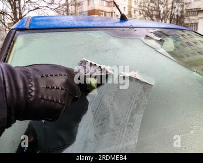 Autofenster im Winter reinigen. Der Mensch kratzt mit einem Plastikkratzer von der Windschutzscheibe eines blauen Autos. Hand in schwarzem Lederhandschuh im C Stockfoto