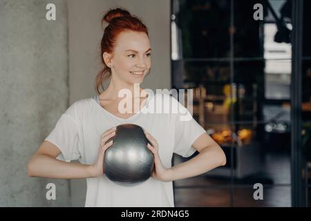 Die sportliche, rothaarige Frau hält einen Sportball und lächelt in einem Fitnessstudio. Bereit für ein Workout. Gesundheit & Pflege. Stockfoto