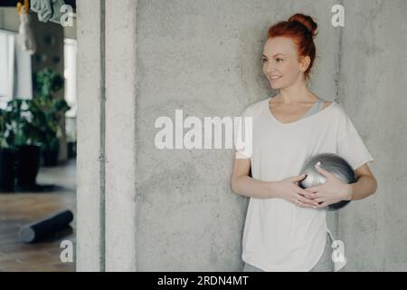 Rothaarige Frau in weißem T-Shirt, hält Fitnessball, steht an der Betonwand des Fitnessstudios und lächelt. Freudige Annahme eines gesunden Lebensstils. Stockfoto