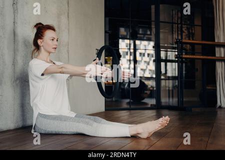 Fokussierte sportliche Frau in weißem T-Shirt und grauen Leggings sitzt im Fitnessstudio und macht Armübungen mit dem Pilates-Ring. Beine ausgestreckt. Stockfoto