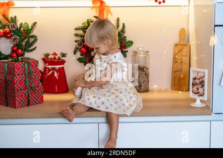 Weihnachtsküche Mädchen. Porträt eines bezaubernden kleinen Mädchens, 2 Jahre alt, sie sitzt auf einem Tisch in einem weißen Kleid und trinkt aus einer roten Tasse in einem dekorierten Stockfoto