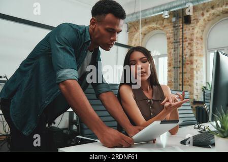 Du musst Folgendes tun. Junge, schöne Frau, die ihrem Kollegen etwas erklärt, während sie in einem modernen Büro arbeitet Stockfoto