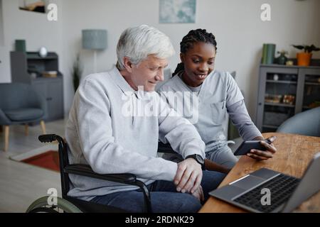Porträt einer lächelnden jungen Krankenschwester, die einem behinderten Mann zu Hause hilft und ein Smartphone zusammen benutzt Stockfoto