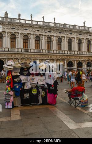 Souvenirstand auf dem Markusplatz, dem Hauptplatz von Venedig, mit der Marciana-Bibliothek und Touristen im Hintergrund im Sommer, Veneto, Italien Stockfoto