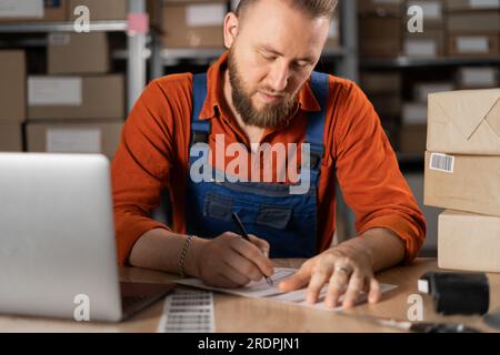 Inhaber eines kleinen Unternehmens, der im Lagerraum mit einem Laptop arbeitet, den Auftrag für den Kunden vorbereitet, ein kleines Paket für die Portoanschrift vorbereitet und die Adresse auf Aufkleber schreibt. Stockfoto