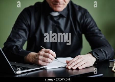 Nahaufnahme eines Senior Priesters, der am Schreibtisch Notizbuch schreibt, während er im Büro an einer Rede arbeitet, im Kopierraum Stockfoto