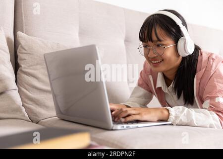 Fokussiertes asiatisches Teenager-Mädchen mit Kopfhörern, das mit Laptop und Buch Notizen schreibt, ein seriöses Highschool-Mädchen. Kleines Mädchen auf der Highschool li Stockfoto