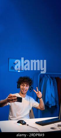 Ein arabischer Gamer sitzt nachts mit blauem Licht am Computer und macht ein Selfie auf einem Smartphone, spielt Videospiele, lächelt und zeigt ein Stockfoto