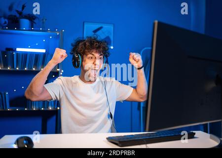 Junger arabischer Gamer, der im Online-Videospiel auf dem Computer gewinnt. Porträt eines Mannes mit Kopfhörern, der beim PvP-Turnier siegte Stockfoto
