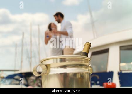 Ein fokussierter Schuss einer geöffneten Champagnerflasche in einem Eiskübel mit einem verschwommenen Hintergrund eines jungen tanzenden Paares auf dem Deck eines Schiffes. Stockfoto