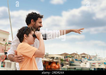 Ein weißer Vater hilft seinem Sohn, ein Fernglas auf einer kleinen Yacht zu benutzen, die am Hafen vor Anker liegt. Sie sehen etwas in der Ferne. Stockfoto