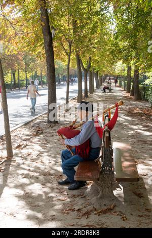 Ein Mann, der Dudelsack spielt und Leute, die im Retiro Park, Madrid, Spanien spazieren gehen. Stockfoto